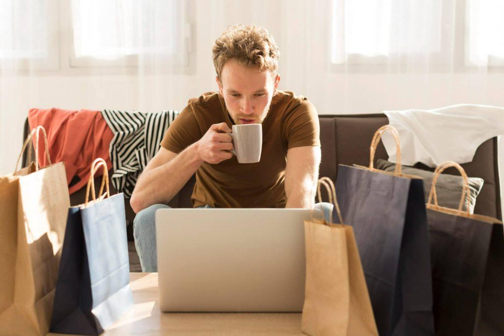 Medium shot van een man die koffie drinkt voor een laptop omringd door boodschappentassen, met de focus op webshop succesvol te verkopen.