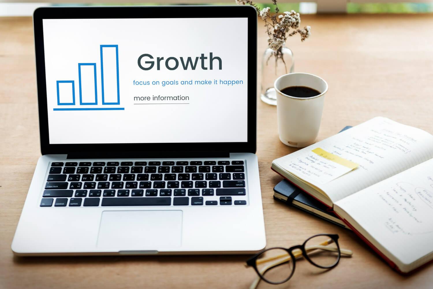 Groei stimuleren: laptopscherm geeft staafdiagram weer met 'groei'-tekst, omringd door bril, notitieboekje en kopje koffie
