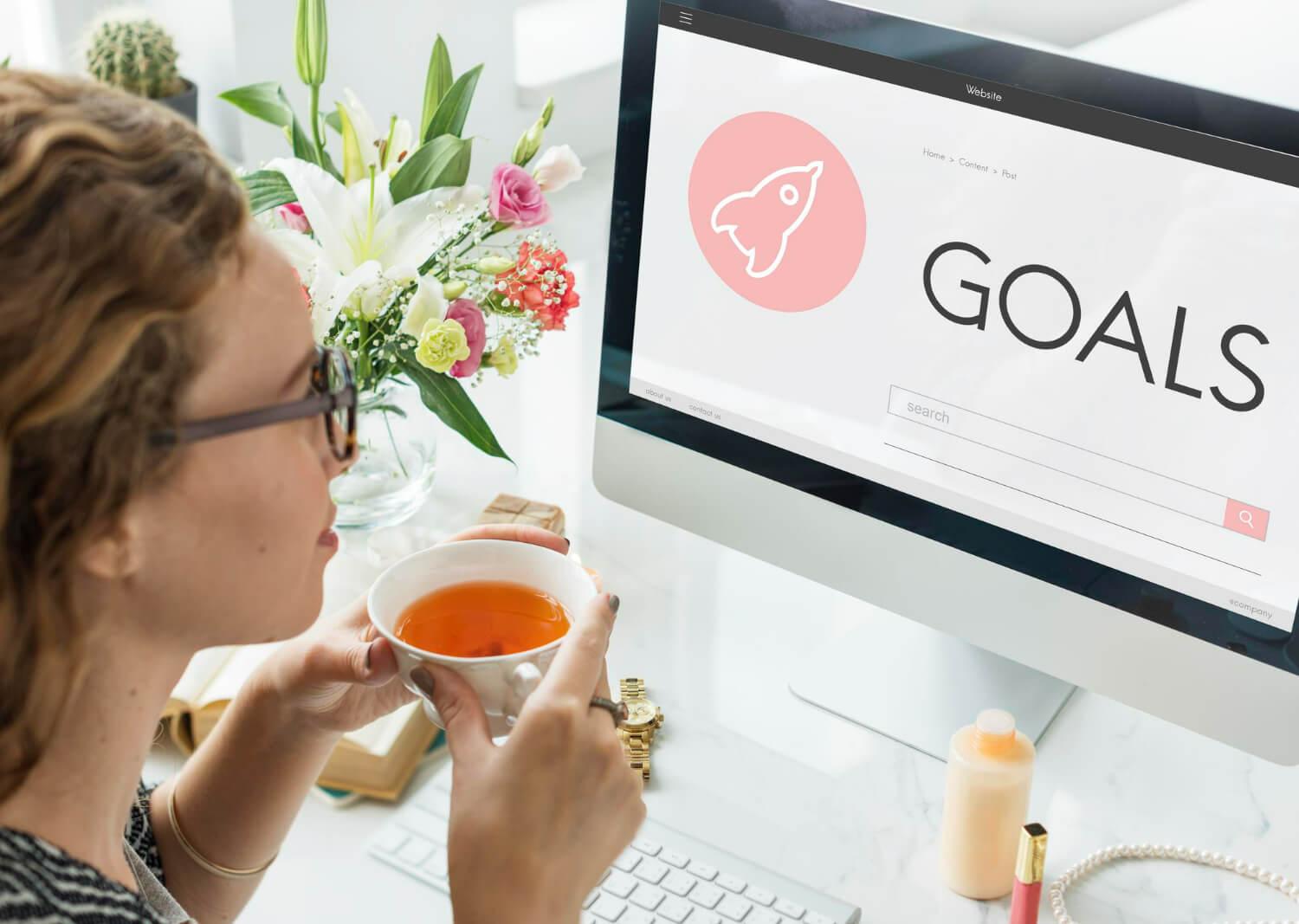 Een dame met een kopje thee starend naar een desktopscherm met een raketsymbool met de tekst 'GOALs' met het ultieme stappenplan voor een webshop