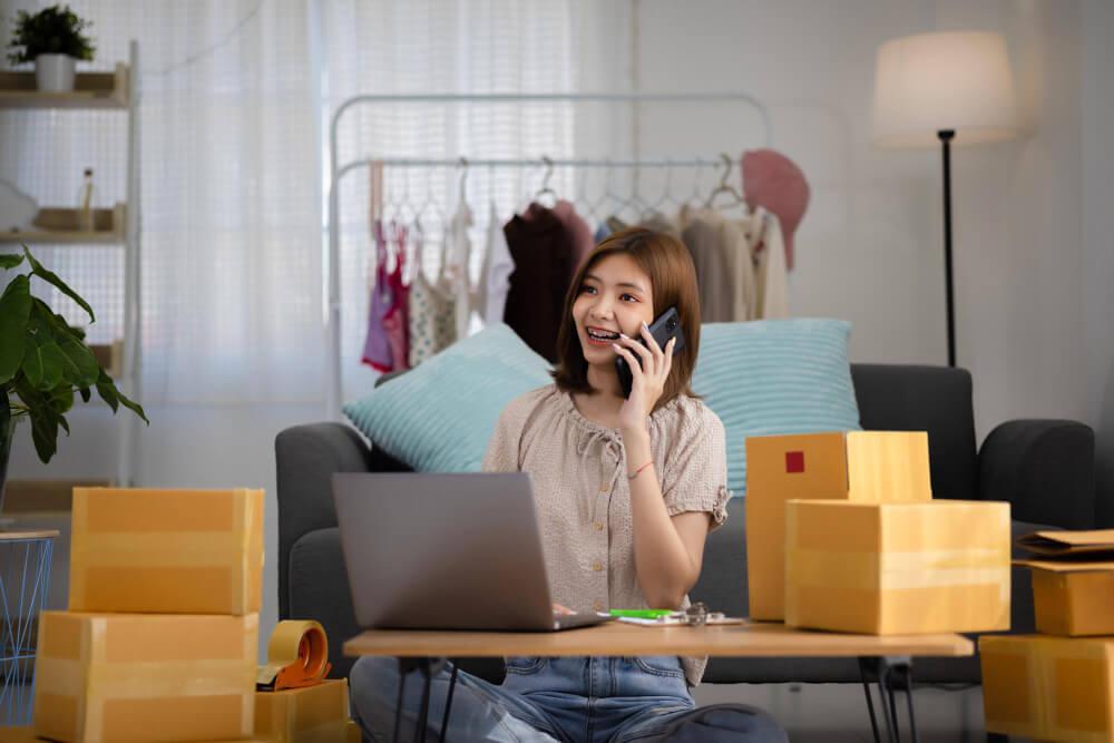 achende jonge Aziatische vrouwelijke ondernemer die telefoontje ontvangt voor nieuwe verkooporder tussen dozen product met laptopcomputer